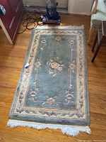 Full view of the rug showing central floral pattern and fringe on edges, placed on a wooden floor next to a vacuum cleaner.