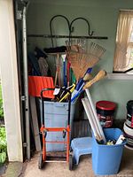 Overall view of hand truck, garden tools such as rakes and brooms in blue bin, shepherd's hooks on wall and paint buckets on floor.
