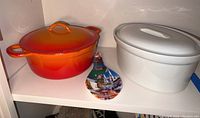 Shelf with Descoware orange-red casserole pot with lid, white Apilco casserole pot with lid, and decorative spoon rest.