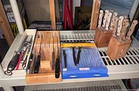 Top-down view of two wooden knife blocks filled with knives, a packaged blue set of disposable cutlery, and a metal peeler on shelving.
