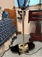 Full view of Beck electric shoe buffer standing upright on carpeted floor next to wooden furniture and bed; shows tall brass-colored metal pole with black knob on top and dual black buffing brushes at base with brass-colored housing.