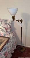 Brass floor lamp next to a floral bed showing the adjustable arm and white glass shade.