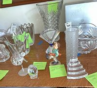 Swarovski crystal ballerina, Goebel figurine, crystal vase, and glass egg on wooden shelf with green labels.