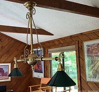 Full view of the brass light fixture hanging from the ceiling with two green glass shades, showing the fixture in a room with wood paneling and framed artwork.