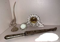 Lot items on shelf showing the quartz mantle clock with decorative glass case, silver-toned letter opener, clear glass bird figurine, and glass ball paperweight.