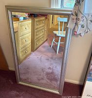 Framed mirror standing against wall showing front full view reflecting furniture and chair.