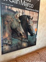 Close-up photo showing the framed exhibition poster featuring bronze horse statues from San Marco, capturing detail and colors of the artwork.