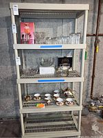 Front view of the plastic shelf unit showing four shelves containing various glassware and china items not included in sale. Shelf unit appears beige with vertical supports and slatted horizontal shelves.
