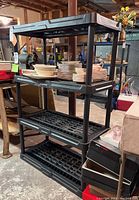 Oblique view of black plastic 3-tier shelf unit with dishes stored on the top shelf in a basement setting.