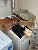 Photo showing seven bags and purses arranged on a white surface including tan leather tote, snakeskin clutch, beige quilted purse with chain, black crocheted bags, cream textured bag, and one covered in clear plastic.