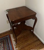 Full view of the square wood side table placed against a corner wall on a hardwood floor, showing the entire structure including legs and lower shelf.