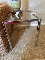 Side table placed next to patterned sofa on carpeted floor, showing glass top and metal legs from a low angle.
