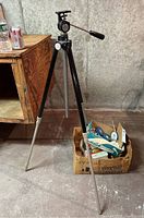 Full view of the Empire tripod with a box of slides and reels on the floor next to it, showing the tripod's metal legs and black head.