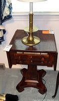 Full front view of a vintage wooden drop leaf side table with a brass lamp on top. Shows table's drawer fronts, pedestal base, and general wood finish.