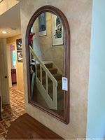 Full view of the framed wall mirror mounted on a beige textured wall, showing an arched top and brown wooden frame. Reflections visible of stairs and hallway.