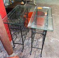 Set of two wrought iron wine rack tables with clear glass tops, showcasing scrollwork frames and circular bottle holders on an indoor concrete floor.