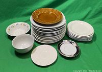 Photo of mixed lot of 25 porcelain plates and 2 bowls stacked on green cloth including brown dinner plates, white embossed plates, floral patterned salad plates, and small decorative plates and bowls.