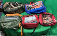 Photo of five bags showing a green and beige duffle, a green and orange fanny pack, a blue NASCAR cooler bag, and two red NASCAR cooler bags with racing graphics and a driver image.