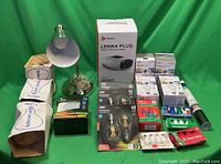 Photo showing entire lot including desk lamp, hand massager box, multiple small boxes of light bulbs and batteries arranged on green background.