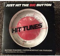 Front view of the original packaging showing the circular radio with a red push button dial labeled 'HIT TUNES' with frequency and volume markings.