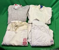 Four fencing jackets, two pairs of fencing pants, and one vest neatly folded together on green backdrop. The jackets show different fabric textures; mesh-like and smooth. Visible All Star brand tags on jackets and pants.