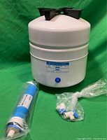 White cylindrical water storage tank with blue outlet valve on green background, plastic fittings visible in front.