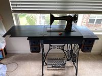 Photo showing the full Singer sewing machine integrated into a wood cabinet with drawers and the machine's decorative black body with gold accents.