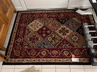 Full view of the approx. 46x65 inch oriental style area rug with multicolored diamond and floral pattern near front entryway with dumbbells on the right side.