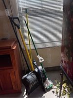 Photo of a yellow snow shovel with D-handle, green-handled snow brush with white bristles and ice scraper, a mop or broom with black handle, and a brown handled broom or mop, standing upright near a window.