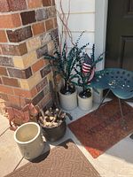 Photo of the tractor seat chair, two ceramic jugs with faux eucalyptus branches and flags, stoneware crock, and cast iron pail with wood positioned by brick corner.