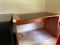 Side view of small wood laminate veneered desk with black pad on surface, positioned on carpeted floor next to dark brown furniture.