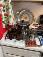 Overall kitchen lot showing two-tier aluminum serving platter, brown coffee cups, candle holders, vintage biscuit tin, and decorative plate.