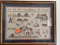 Framed cross stitch farm scene showing rural landscape with people, animals, and buildings, bordered, with text along the top edge.