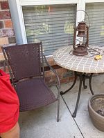 Round outdoor tile top table with metal legs next to a woven metal frame chair and window background