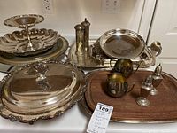 Wide view of silver plated serving pieces including platters, pedestal dish, covered chafing dish, salt and pepper shakers, sugar, creamer, and tray.