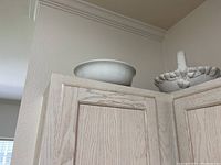 White ceramic bowl and white ceramic strawberry basket sitting on top of a kitchen cabinet. The basket has a handle and strawberry decorations.