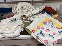 Photo showing assorted folded table linens on a kitchen counter, including embroidered napkins, patterned napkins with strawberries, and place mats.
