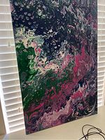 Abstract wall art with swirling green, pink, black, and purple colors shown in natural light against blinds.