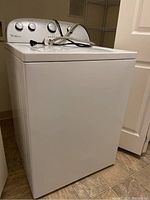 Front and side view of white Whirlpool top loading washing machine in a laundry area.
