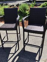 Front view of two black woven bar stools with armrests, showing seat height and woven texture.