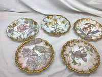 Five hand-painted porcelain plates with gold scalloped edges displayed on fabric background, showing front floral designs.