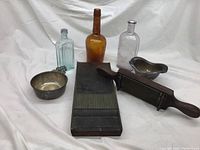 Full lot showing all items on white fabric backdrop: sterling silver bowl with handle, silver miniature bathtub, three medicine bottles in amber, clear and light blue glass, and wooden antique pill maker.