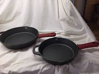 Two black cast iron frying pans on white background. One is larger with double handles and both have red silicone sleeved handles.