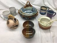 Photo showing entire pottery lot with mugs, covered serving dish, small soup tureen, two small bowls, and decorative pitcher