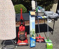 Full view of cleaning lot items including ironing board, Hoover vacuum, cleaning pole, dustpan and broom, mop, and cleaning supplies on outdoor pavement and table.