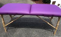 Full view of purple padded massage table on pine wooden frame outdoors.