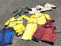 A group photo of outdoor jackets and rainwear laid on pavement showing a variety of colors and styles including yellow rain suits, puffer vest, and insulated jackets.