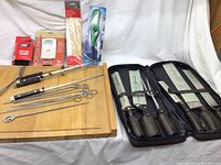 Full display of cutlery set in leather case, grilling metal skewers, cutting board, and Weber grilling accessories in original packaging spread on a white background.