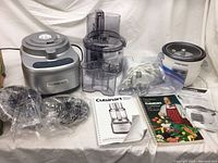 Full lot view showing all items on white tablecloth: food processor base, accessories and blades, rice cooker, hand mixer, manuals and recipe booklets.