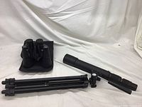 Photo showing black spotting scope, tripod, and binoculars arranged on white fabric background